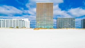 A beachfront hotel with multiple floors, a sandy beach in the foreground, and a clear blue sky with clouds. - New Complex!  Lazy River/Slide, Beachfront! (Orange Beach)