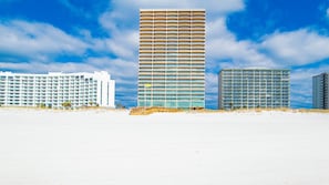 Un hotel frente a la playa con varios pisos, una playa arenosa en primer plano y un cielo azul despejado.