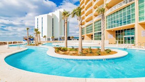 A pool area with palm trees, a white building, and a sign indicating a lazy river. - New Complex!  Lazy River/Slide, Beachfront! (Orange Beach)
