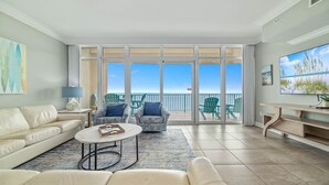 A spacious living room with a large sliding glass door leading to a balcony with a view of the ocean. There are two patterned armchairs, a white sofa, and a coffee table with a magazine. - New Complex!  Lazy River/Slide, Beachfront! (Orange Beach)