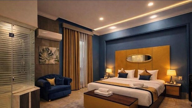 A hotel room with a large bed, a blue armchair, a bedside table with a lamp, and a large mirror on the headboard. - GA PLUS (Cairo)