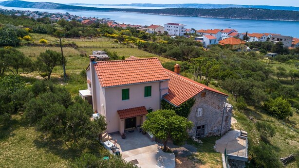 Une maison à deux étages, avec un toit recouvert de tuiles rouges, entourée de verdure et offrant une vue sur une ville côtière.