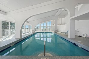 En indendørs swimmingpool med klart, blåt vand, hvide vægge og store vinduer.