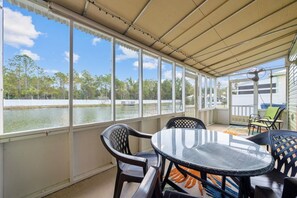 Un espacio al aire libre techado con una mesa y sillas, ventanas amplias con vista a un cuerpo de agua y árboles.