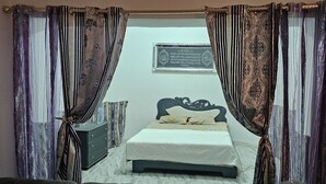 A bedroom with a bed, curtains, a nightstand, a water bottle, and a framed item on the wall. - Villa Atlas Osis (As-Sabikah)