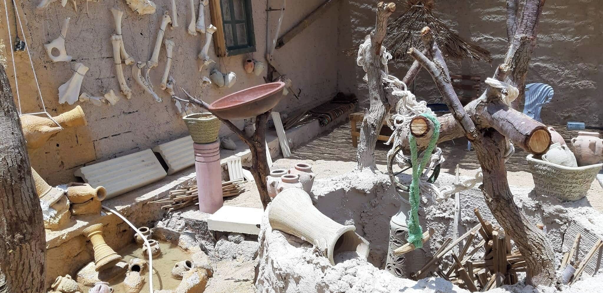 Un décor extérieur rustique agrémenté de divers objets en céramique et en bois, notamment un bassin rose, des tours de potier et un arbre avec une balançoire en corde.