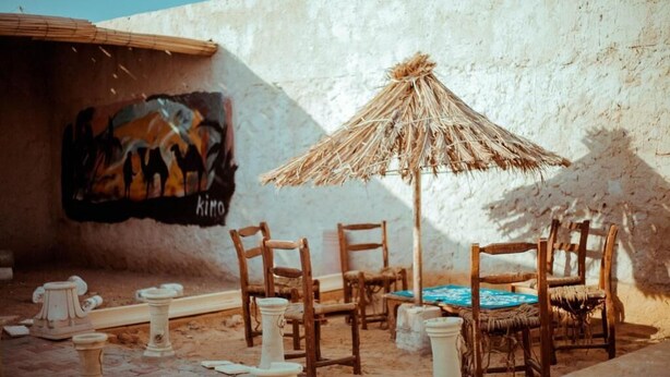 A rustic outdoor seating area with wooden chairs and a straw umbrella, a mural on the wall, and a stone bench. - Villa Atlas Osis (As-Sabikah)