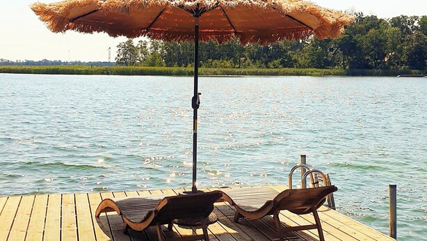A wooden dock with a thatched umbrella and two wooden chairs.