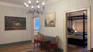 Una habitación con piso de madera, un candelabro, una cama con dosel, una pequeña mesa con sillas y dos cuadros en la pared.