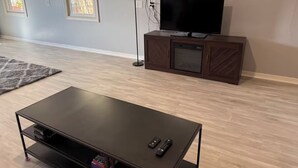 Una sala de estar con un televisor de pantalla plana sobre un mueble de madera, una mesa de centro negra y una lámpara de pie.