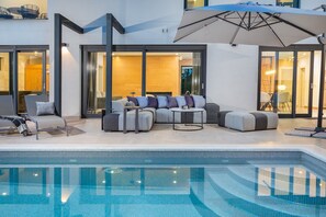 A modern outdoor pool area with a sectional sofa and lounge chairs, a small round table, and a large umbrella.