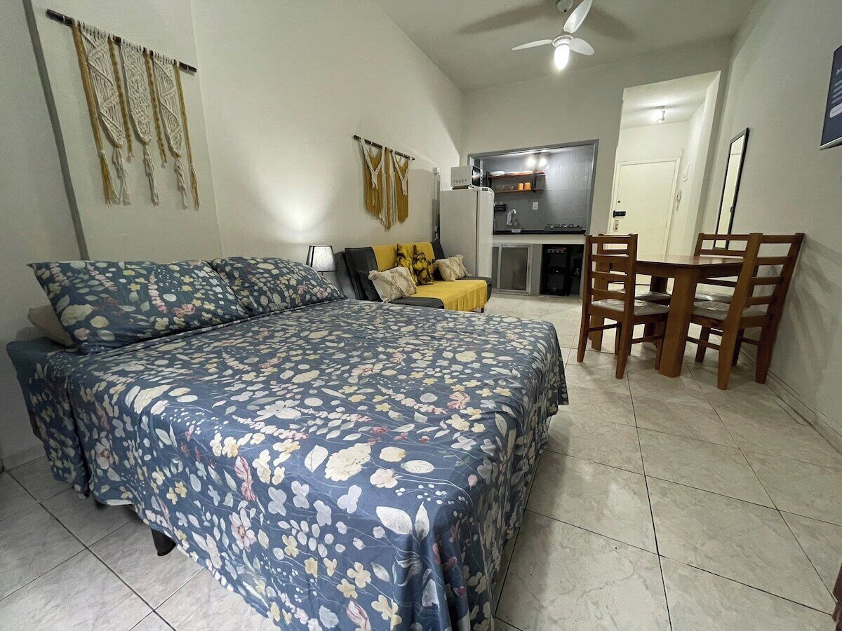 A bedroom with a bed, a chair, a small table, and a kitchenette in the background.