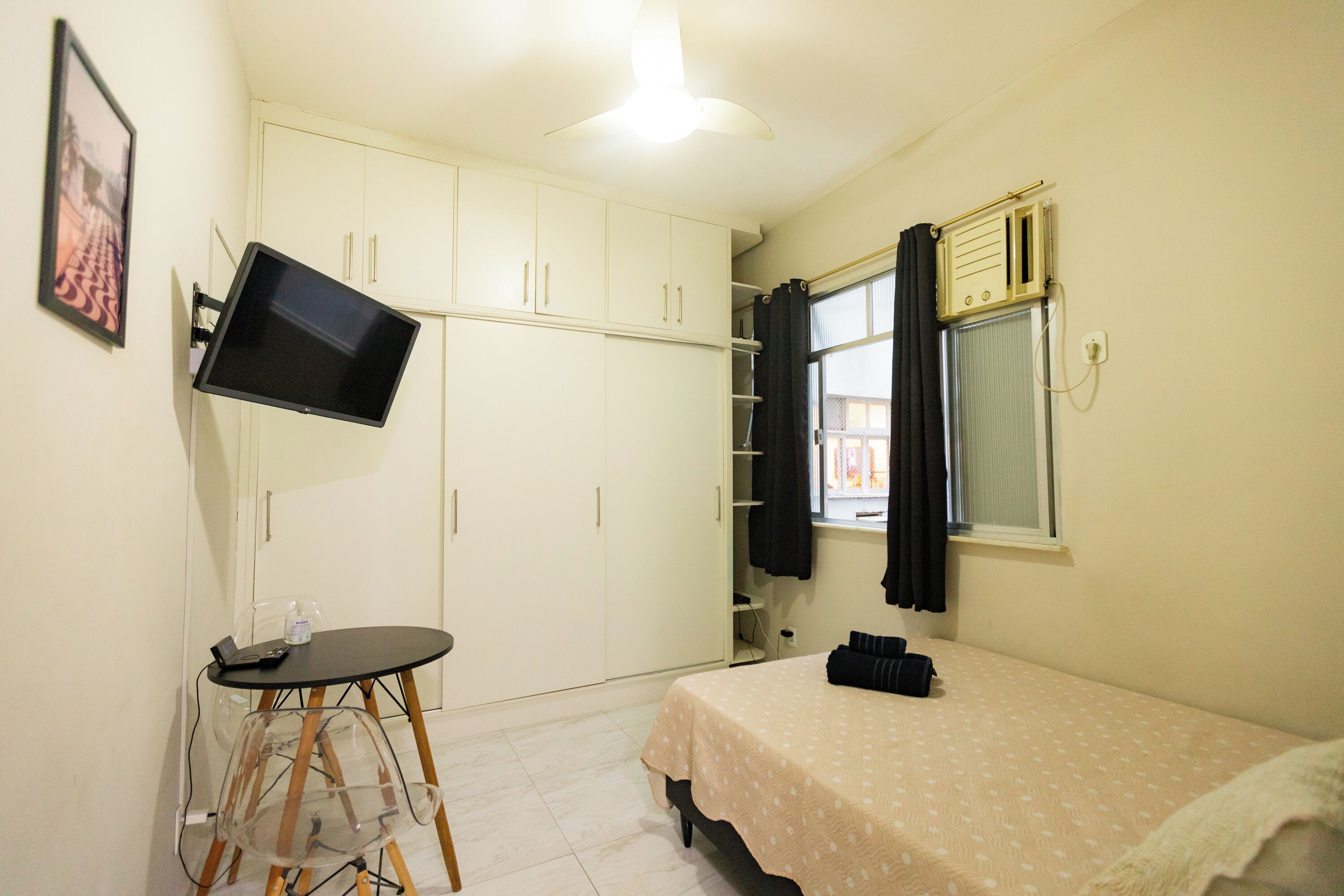 A small bedroom with a bed, a desk with a chair, a television mounted on the wall, and a wardrobe.