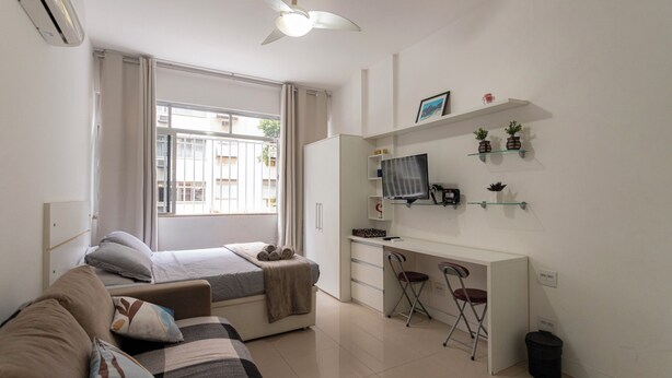 Camera d'albergo compatta con un letto, una scrivania e una sedia. Si vede una finestra con tende, una ventola a soffitto e un piccolo cestino.