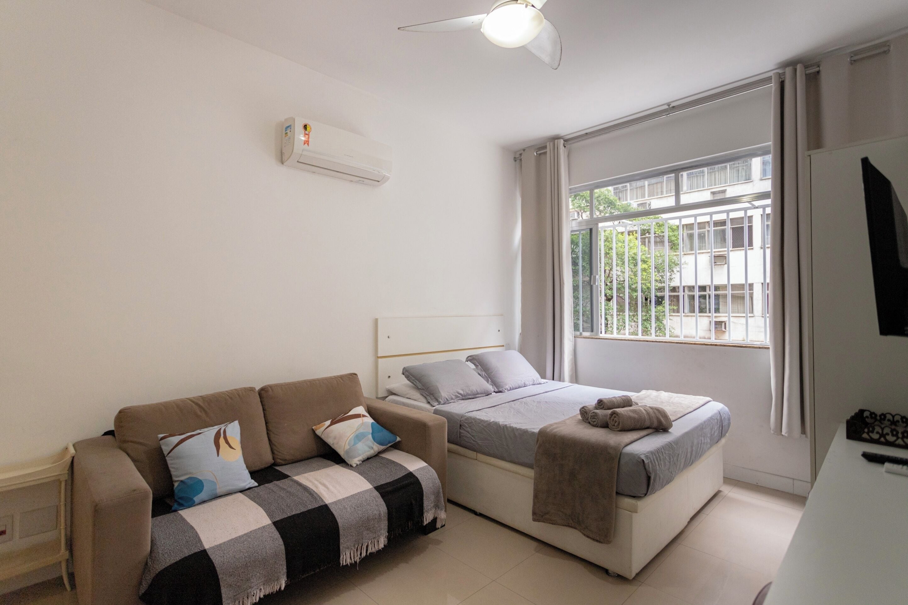 Un dormitorio con cama, sofá, escritorio y ventana con vista a edificios.