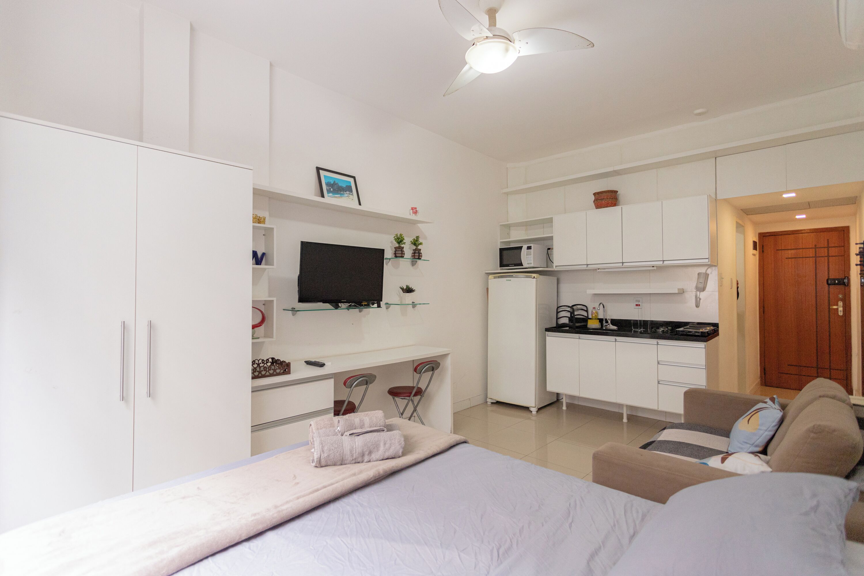 A modern, well-lit room with a bed, a TV, a kitchenette, and a wooden door.