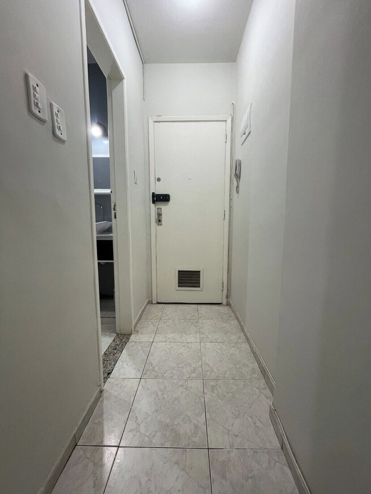 Un corridoio stretto con una porta bianca in fondo, pavimento piastrellato e interruttore della luce al muro.