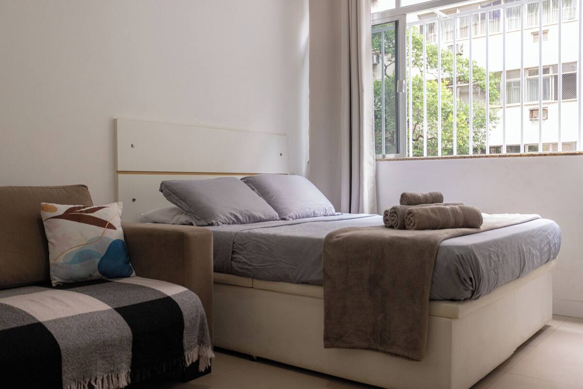 A bedroom with a bed, a sofa, and a window with a view of buildings.