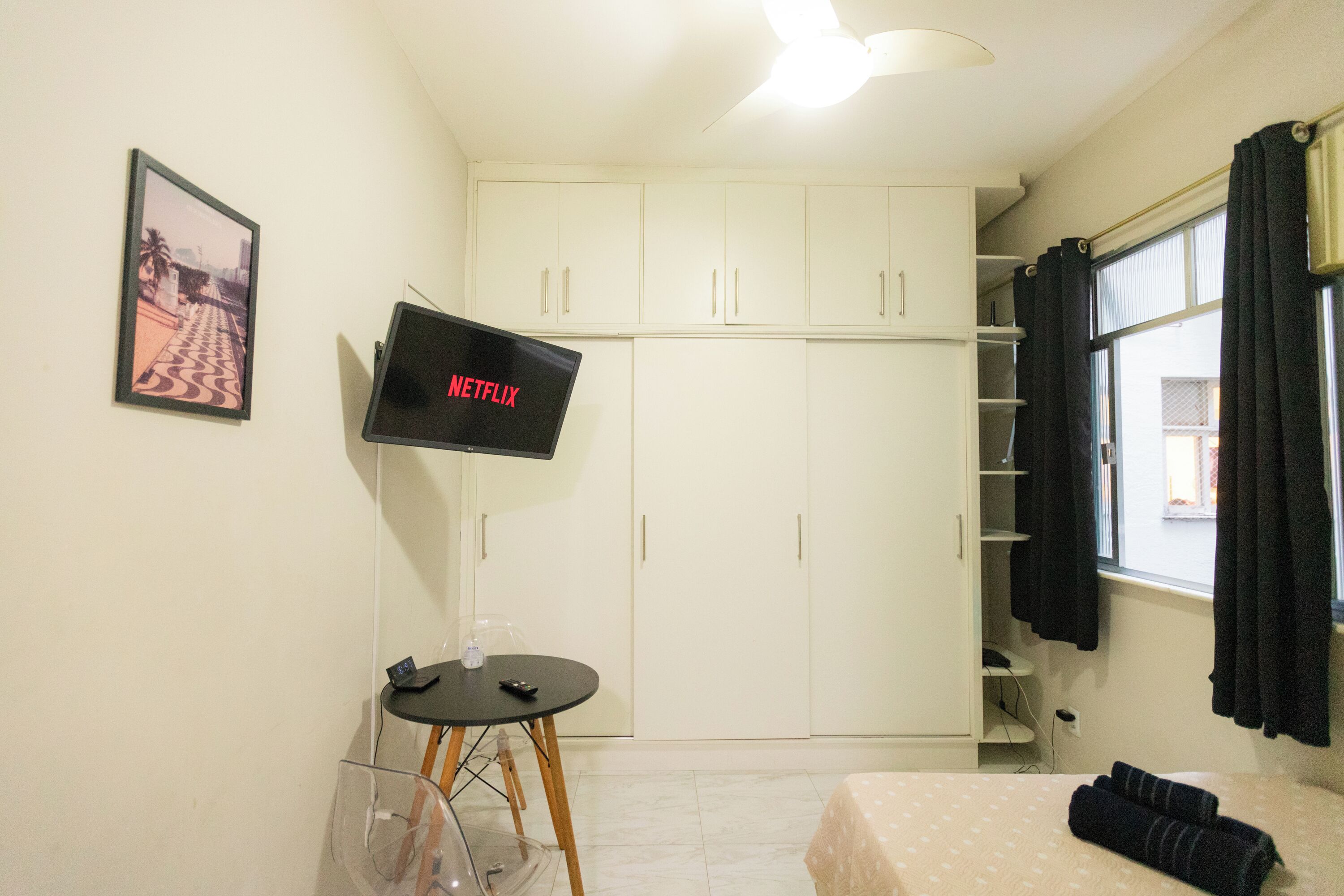 Una habitación pequeña con una cama, un televisor montado en la pared, una mesita con una botella y un control remoto, y un armario con espejo.