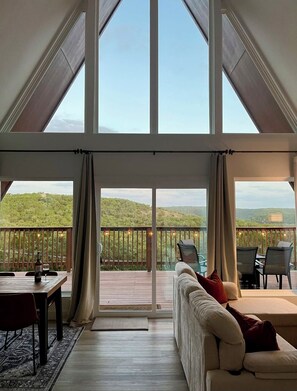 A spacious room with a large window, a wooden deck, a sofa, and a dining table. - Cozy Hillside A-Frame / Hill Country Views / Close to Lake & River (Canyon Lake)