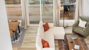A spacious loft with a high ceiling, large windows offering a view of a lush landscape, a white sectional sofa, a green armchair, and a wooden coffee table.