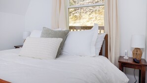 A neatly made bed with white bedding and several pillows, a bedside table with a lamp and a remote control, and a window with curtains.