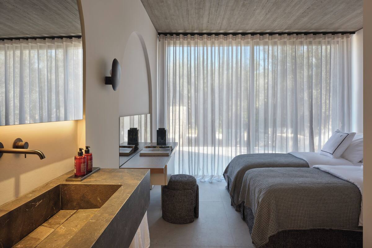 A modern hotel room with a large bathroom sink, a mirror, a bedside table with toiletries, two beds with checkered bedding, and sheer curtains.