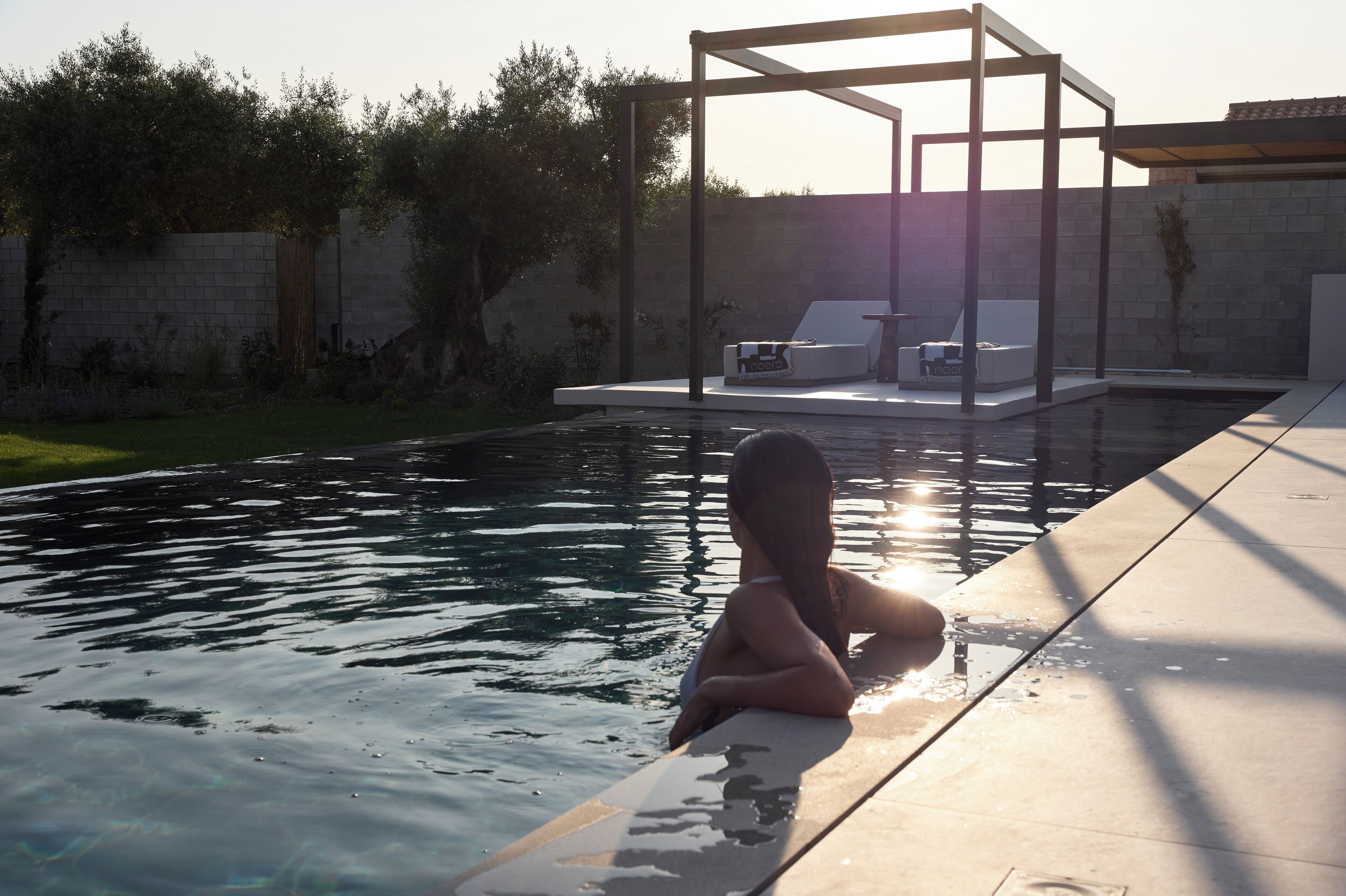A swimming pool with a sunlit deck area, a pergola structure, and a lounge area with chairs and a table.