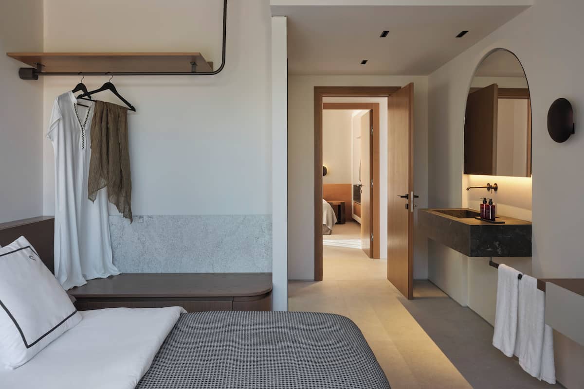 A modern hotel room with a bed, a bench, a hanging clothes rack, a bathroom with a mirror and sink, and a hallway leading to another room.