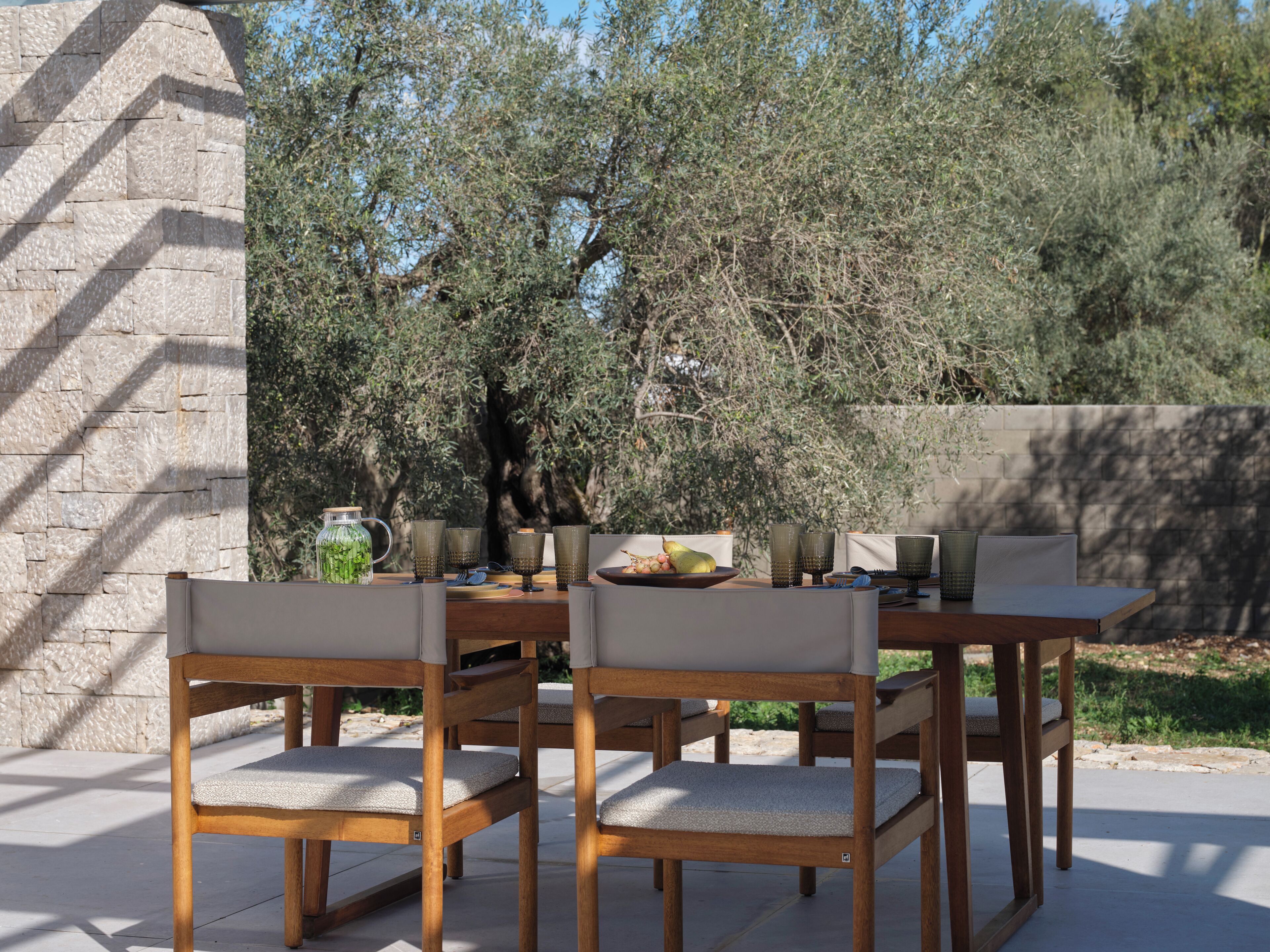 An outdoor dining area with a stone wall, wooden chairs, and a table set with glassware and a bowl of fruit.