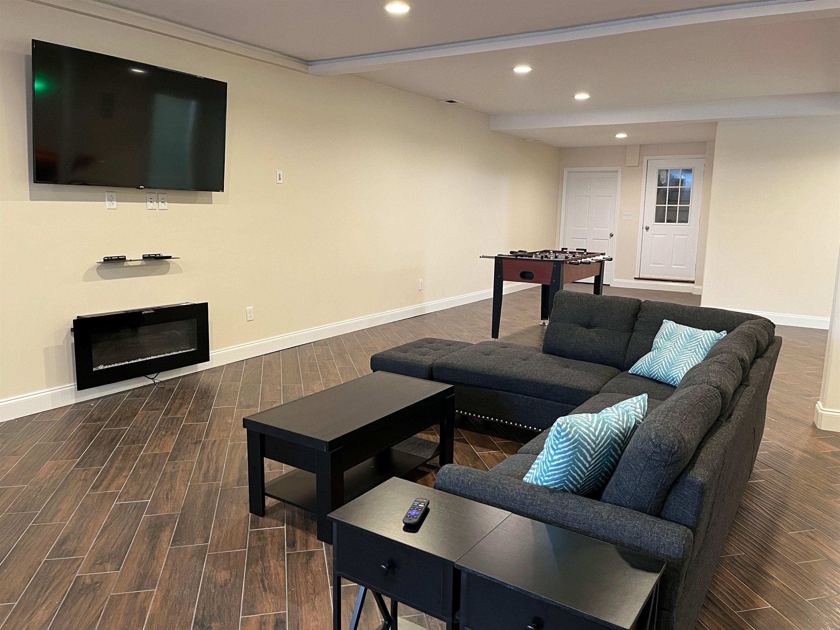 A modern living room with a sectional sofa, a flat-screen TV mounted on the wall, a foosball table, and a fireplace.