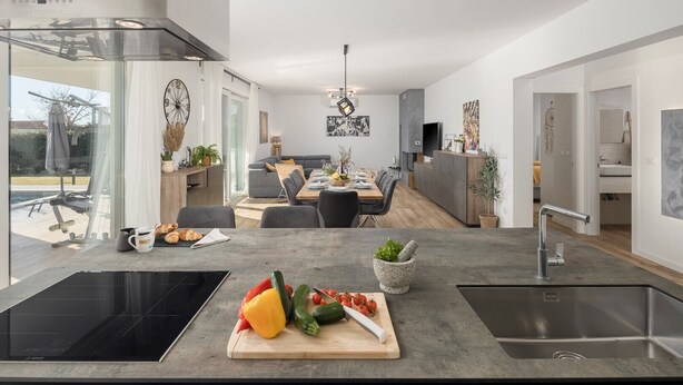 Una cocina moderna con una isla central, un área de comedor con sofá gris y mesa de madera, y vistas al exterior.