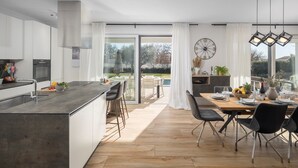 Una cocina moderna con una isla central, comedor con mesa de madera y vistas a un jardín a través de ventanas amplias.