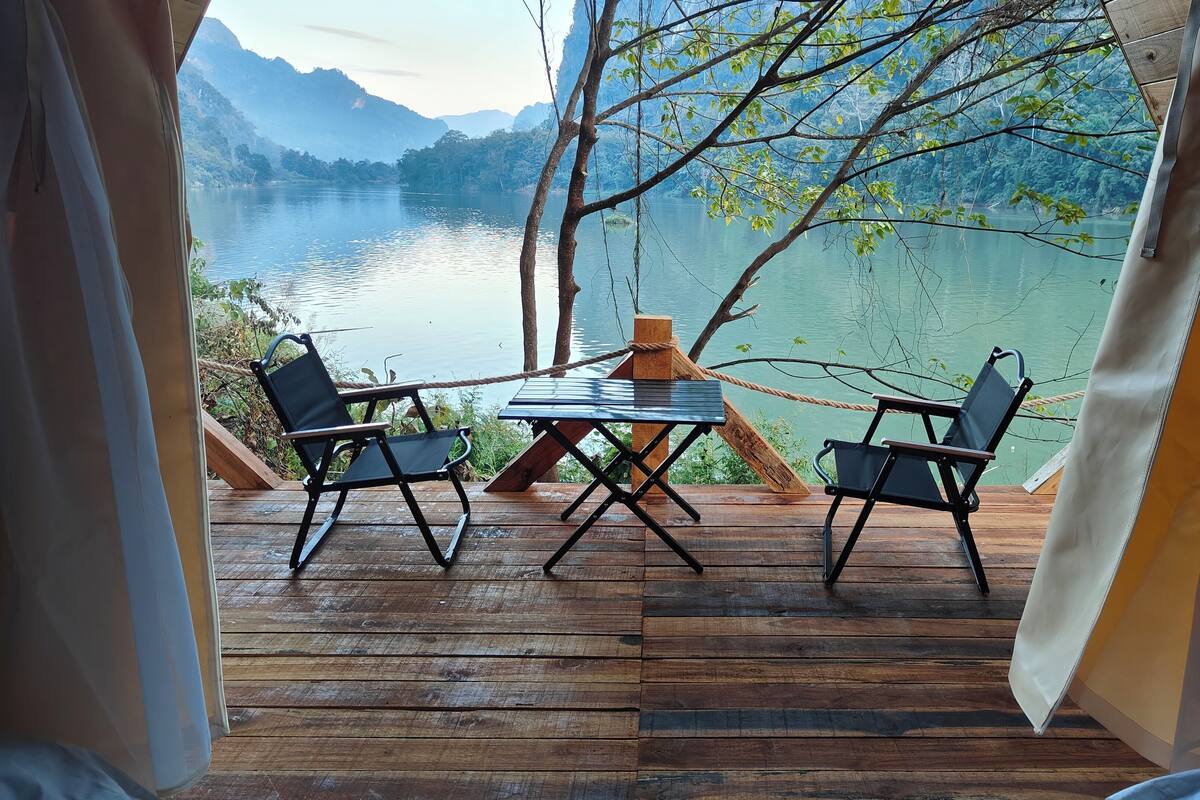 Eine Holzterrasse mit Tisch und zwei Stühlen mit Blick auf einen See, im Hintergrund Berge.