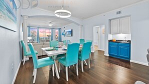 A modern dining area with teal chairs and a white table, a kitchen with blue cabinets, and a living room with a large window.
