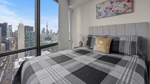 A bedroom with a large window, a city view, a bed with a checkered blanket, and a floral painting on the wall. - GLOBALSTAY Magnificent 1 Bedroom High Floor View (Toronto/Canada)