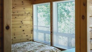 A bedroom with wooden walls, a bed with a floral bedspread, and two windows with blinds. - Pike's Place Cabin - Lakewood Park Cabins (Kenora, Unorganized)