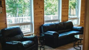 A room with a black leather sofa and armchair, a small round table, and large windows with a view of a wooden deck and trees. - Pike's Place Cabin - Lakewood Park Cabins (Kenora, Unorganized)