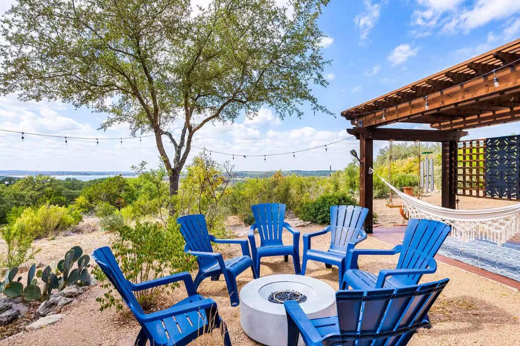 A patio area with blue Adirondack chairs arranged around a circular fire pit, overlooking a scenic landscape with trees and a distant view.