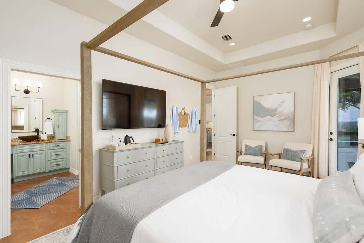 A bedroom with a large bed, a television mounted on the wall, a bathroom with a sink and mirror, and a seating area with chairs.