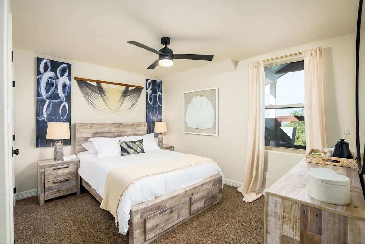 Une chambre à coucher avec un lit en bois, un ventilateur de plafond, une fenêtre avec des rideaux et des œuvres d’art accrochées aux murs.