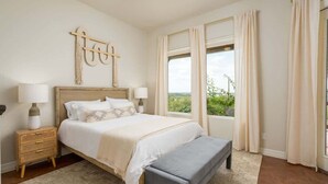 A bedroom with a large bed, bedside tables, a bench, and a view of greenery. - Lake Views, Fire Pit, BBQ & Patio | Pet-Friendly! (Canyon Lake)