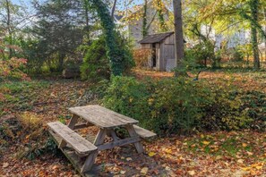A wooden picnic table in a garden with autumn leaves and a small shed in the background.
