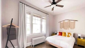 Un dormitorio con cama, radiador, ventilador de techo, ventana con cortinas, piso de madera y mesita de noche con lámpara.