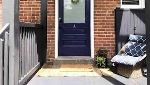Una casa con puerta azul oscuro, fachada de ladrillo, maceta colgante y silla de mimbre con cojines.