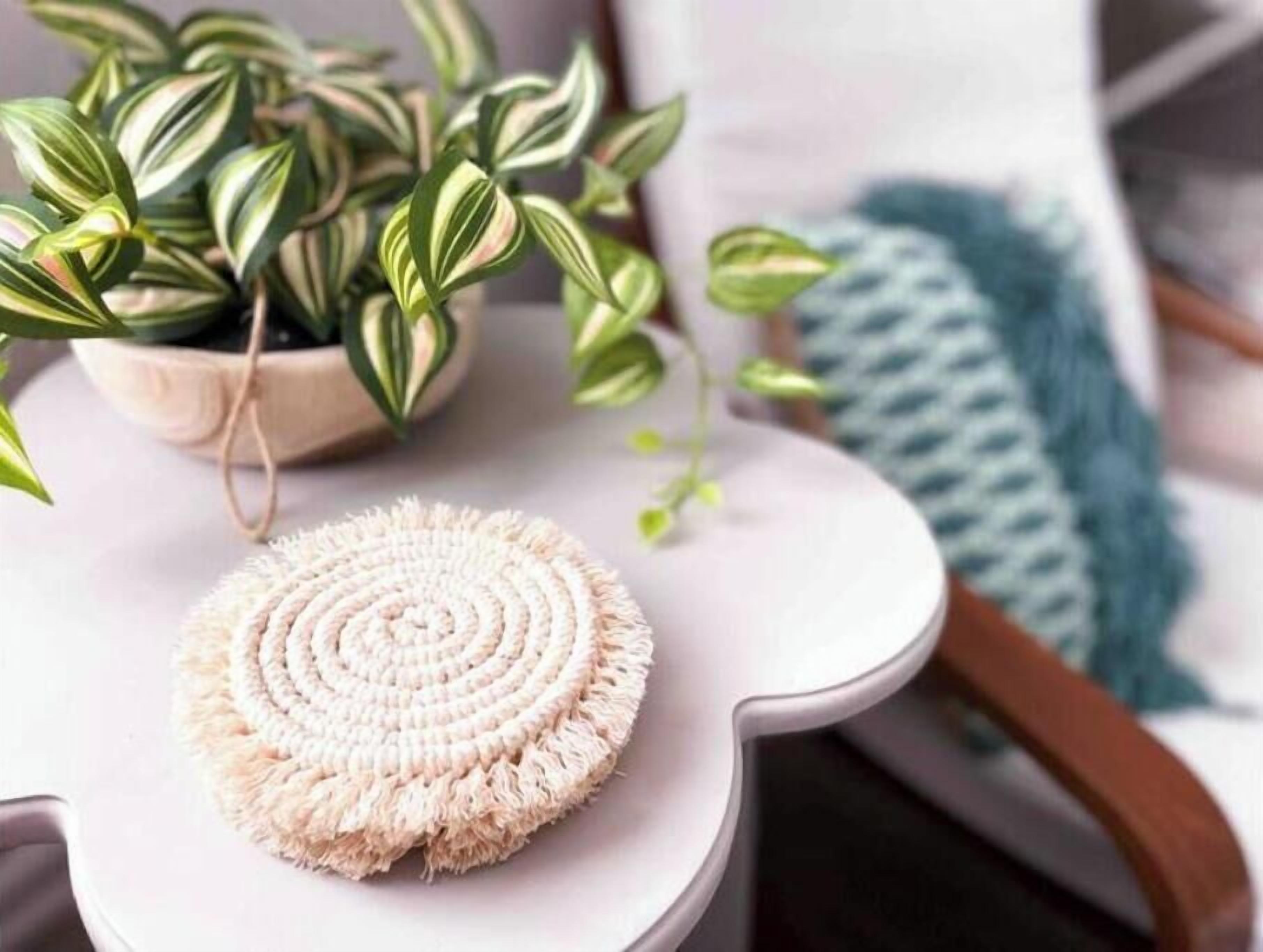 A round, textured mat on a white table with a plant and a blue knitted item in the background.
