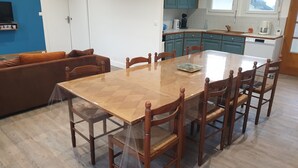 Een eetgedeelte met een houten tafel en stoelen, een keuken op de achtergrond en een blauwe wand met een televisie.