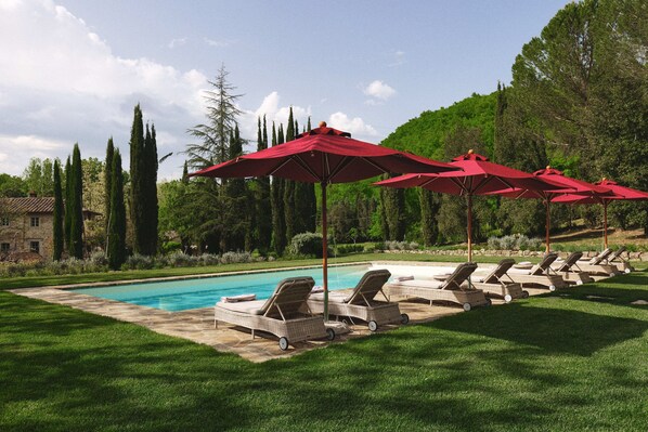 Zona de piscina con sillones de descanso y sombrillas rojas, rodeada de vegetación y árboles.