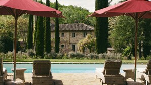 A poolside area with wicker chairs and red umbrellas, a stone building in the background, and a well-maintained lawn. - Villa Mandorla by My Villa Collection (Barberino Tavarnelle)