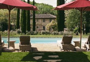 A poolside area with wicker chairs and red umbrellas, a stone building in the background, and a well-maintained lawn.